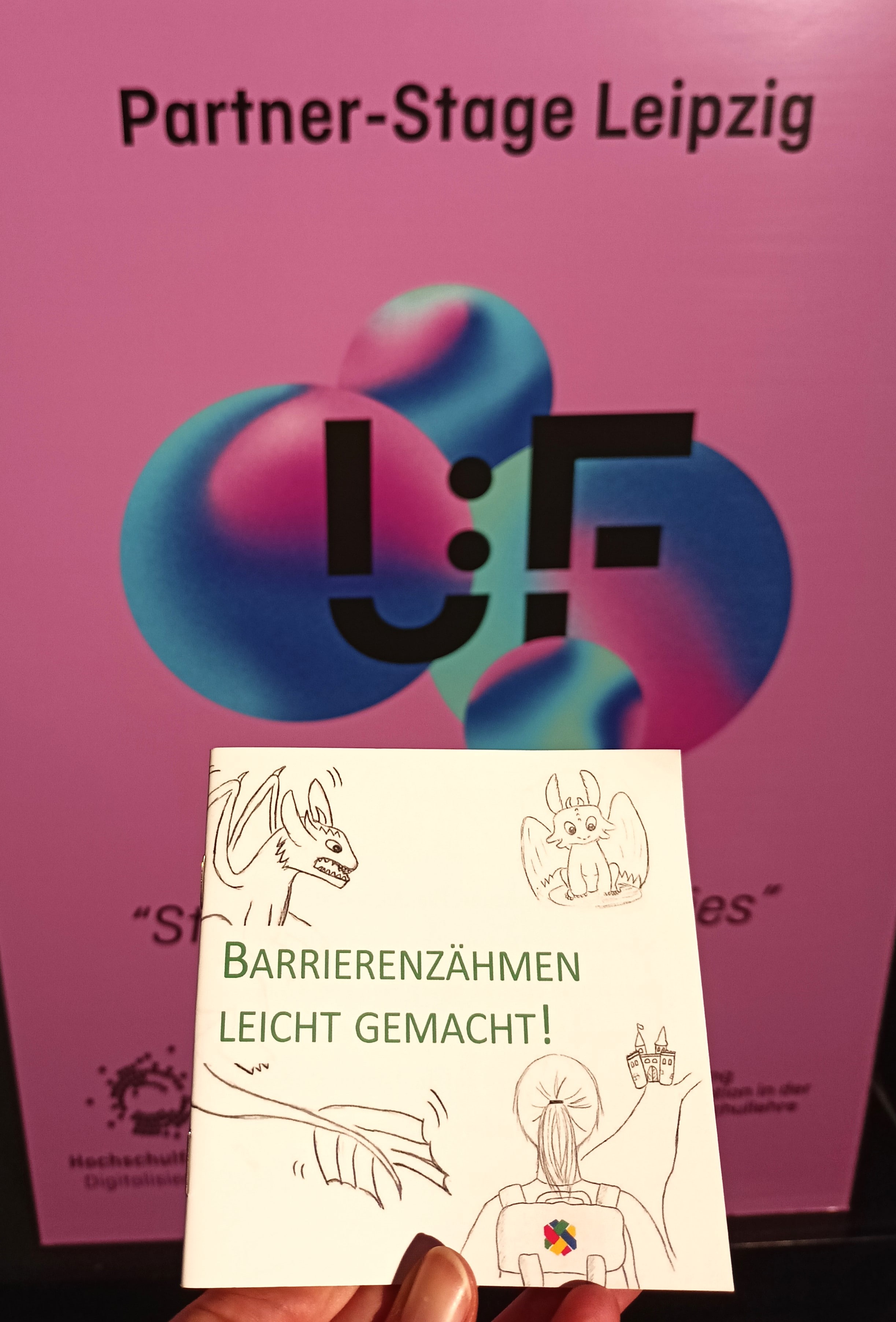 Im Vordergrund hebt eine Hand ein kleines Bilderbuch in die Kamera, auf dem 'Barrierenzähmen leicht gemacht!' steht, sowie Zeichnungen von Drachen und einer Frau mit einem SHUFFLE-Rucksack abgebildet sind. Hinter dem Bilderbuch und etwas außerhalb des Kamerafokus, ist ein UFF-Plakat der Partnerbühne Leipzig zu sehen.