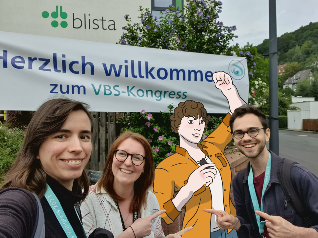 Drei Mitglieder des SHUFFLE-Teams posieren vor einem Banner mit der Aufschrift 'Herzlich willkommen zum VBS-Kongress'. Das gemalte Bild der Figur Gabriel ist in realistischer Größe inmitten der Personen eingefügt.